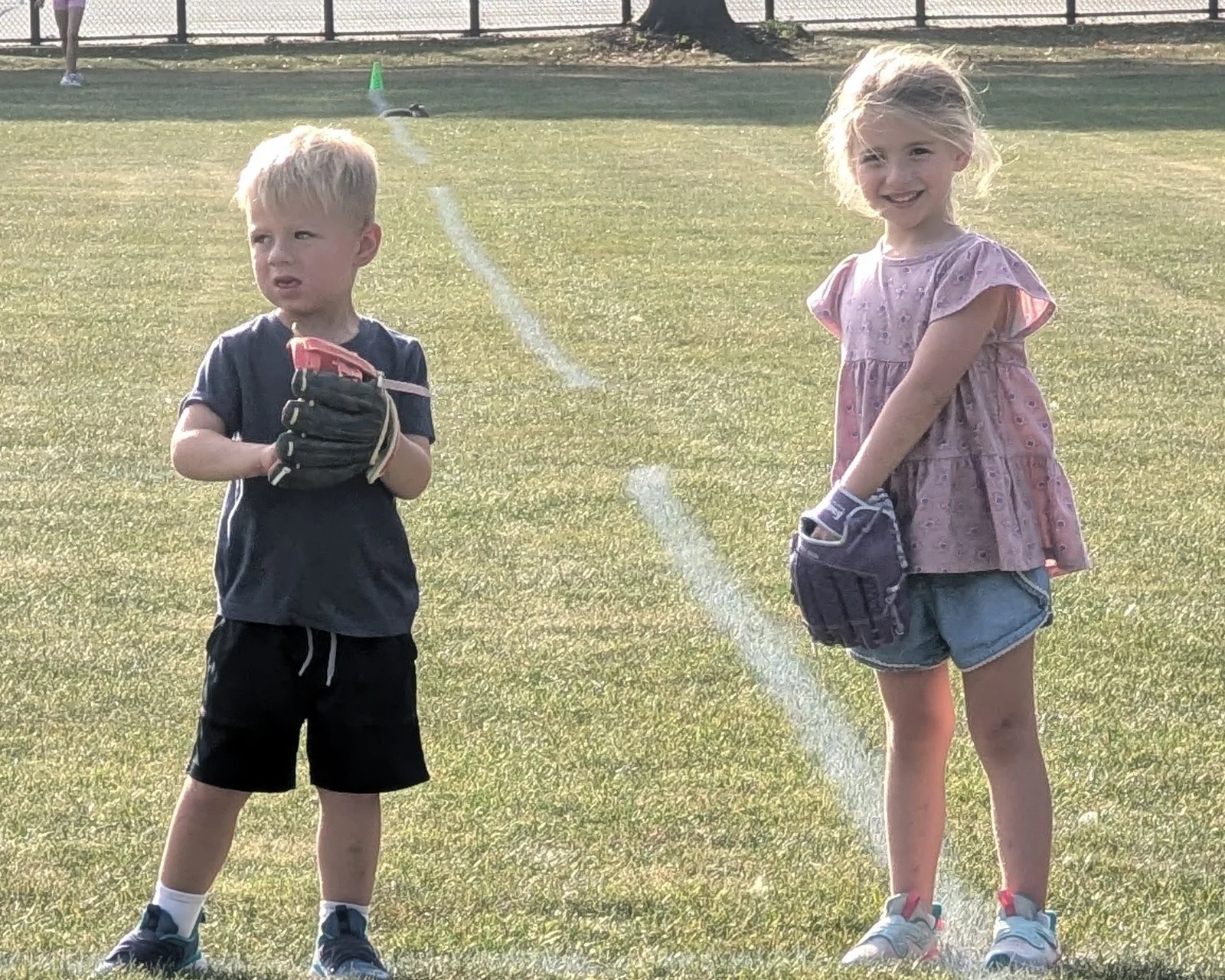 Caleb in a batting helmet and Abigail with a glove, standing together on a sunny T-ball field